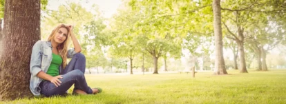 Full length of smiling woman with hand in hair while sitting under tree in park